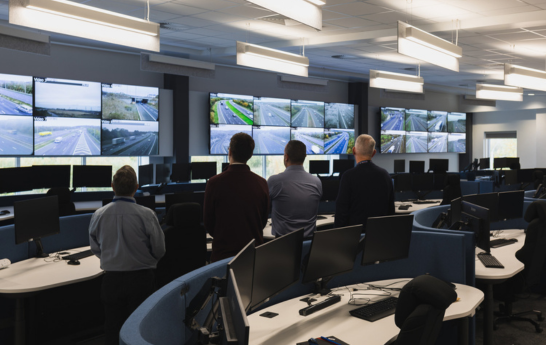 Four men looking at 22 screens on a wall in the control room